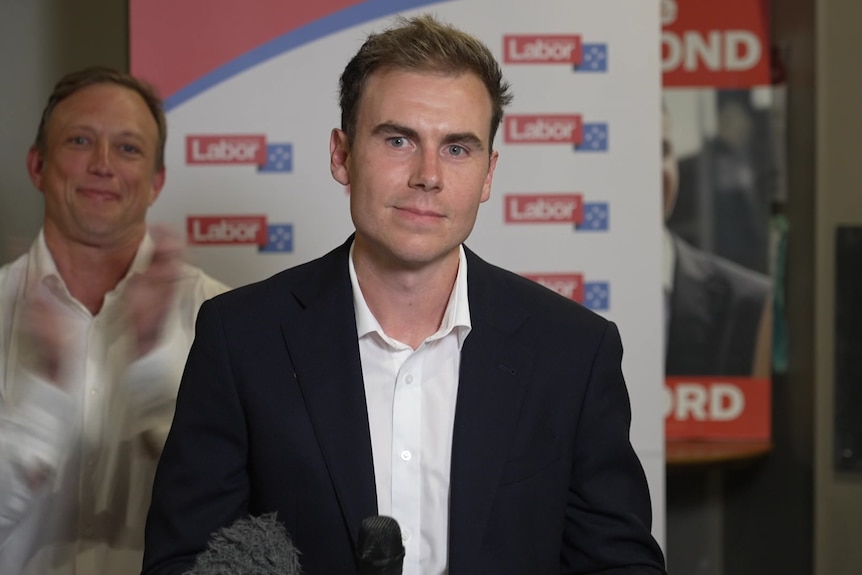 A man in a suit smiling at a press conference with another man in white shirt applauding behind him.