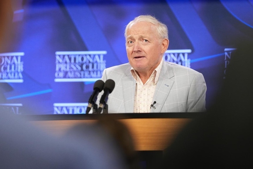 An older man in a grey suit and thinning white hair stands at the National Press Club podium.