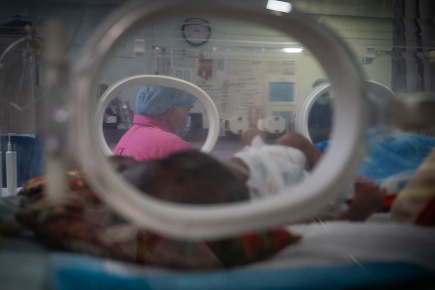 A close up of the conjoined twins in a small crib being overseen by a nurse.