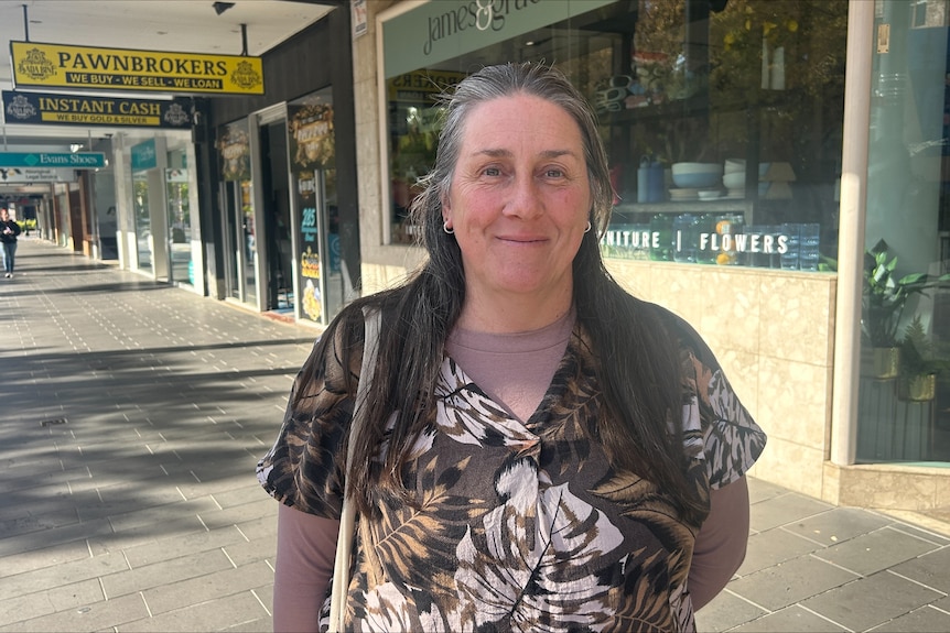 a lady with greying hair smiles at a camera in front of a shopping strip