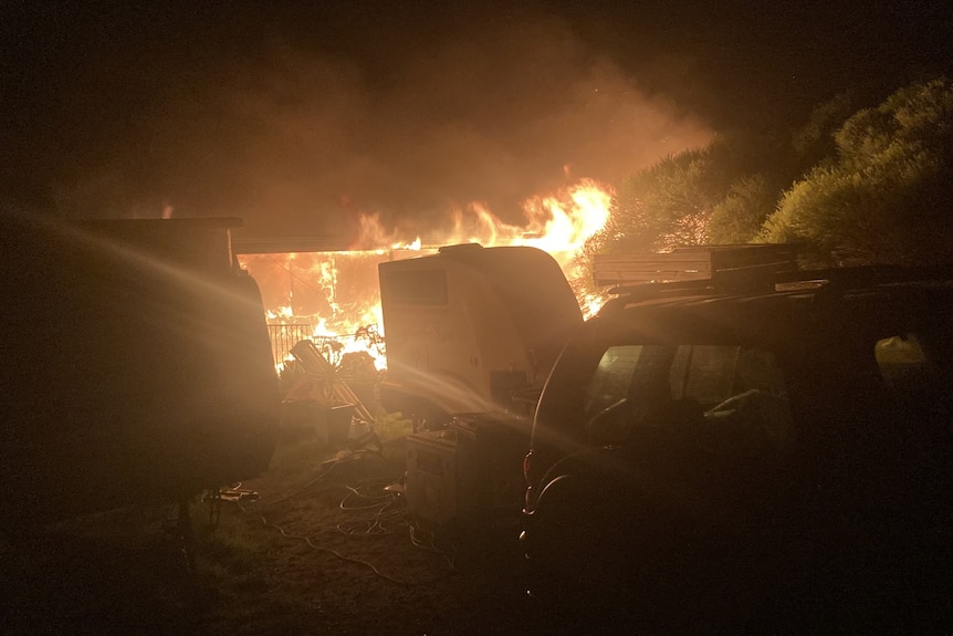 A house on fire with a caravan and car in front of it.