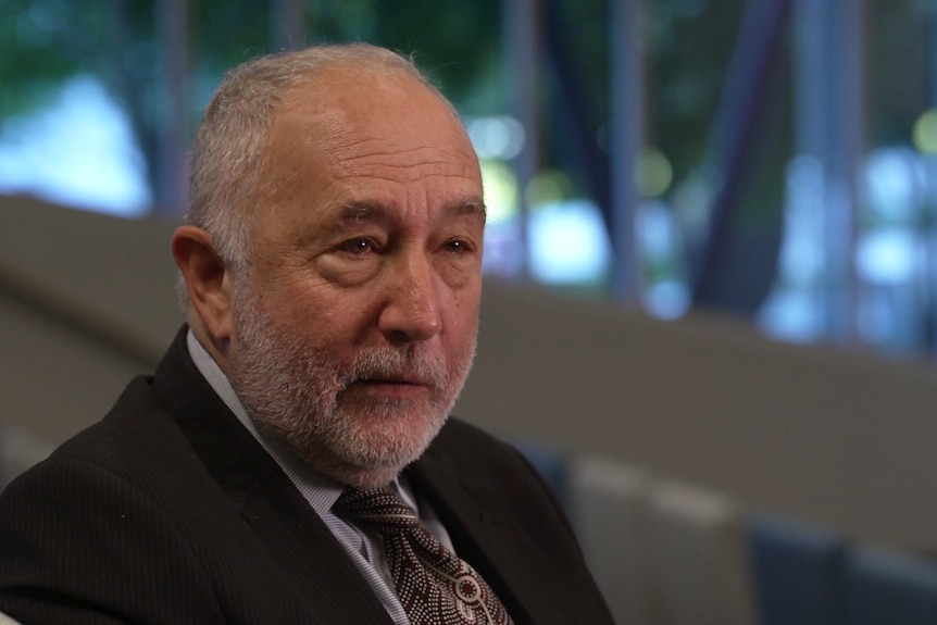 An older man with short grey hair and a grey beard wearing a suit and tie sitting in an empty lecture theatre. 