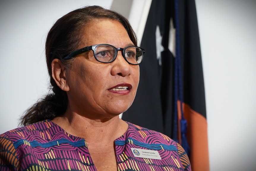 An Aboriginal woman, tanned, dark brown hair, black-framed glasses, purple and blue pattern top standing in front of NT flag