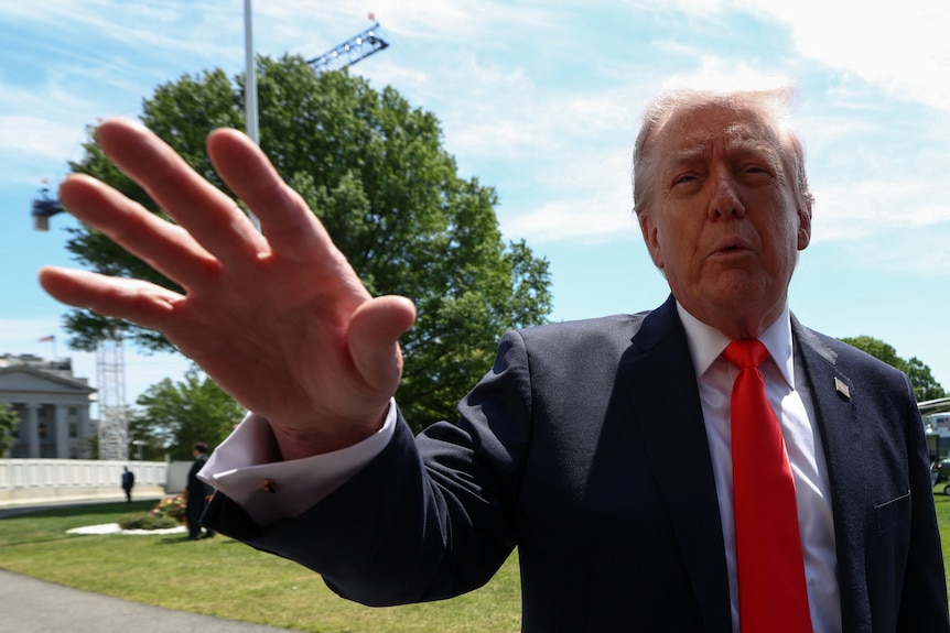 Donal Trump speaking outdoors, with his hand outstretched, palm down.