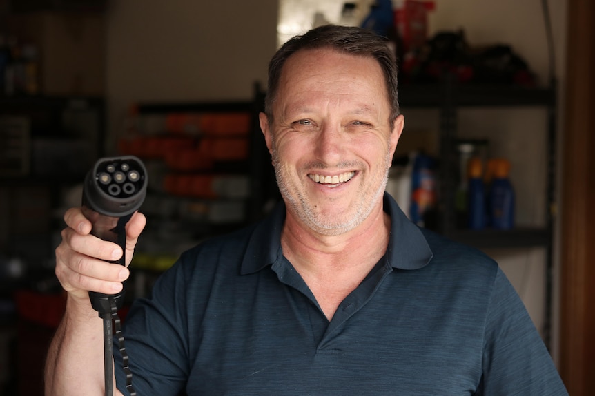 Robin Sands wears a blue polo shirt and holds an EV charger up near his face, smiling.
