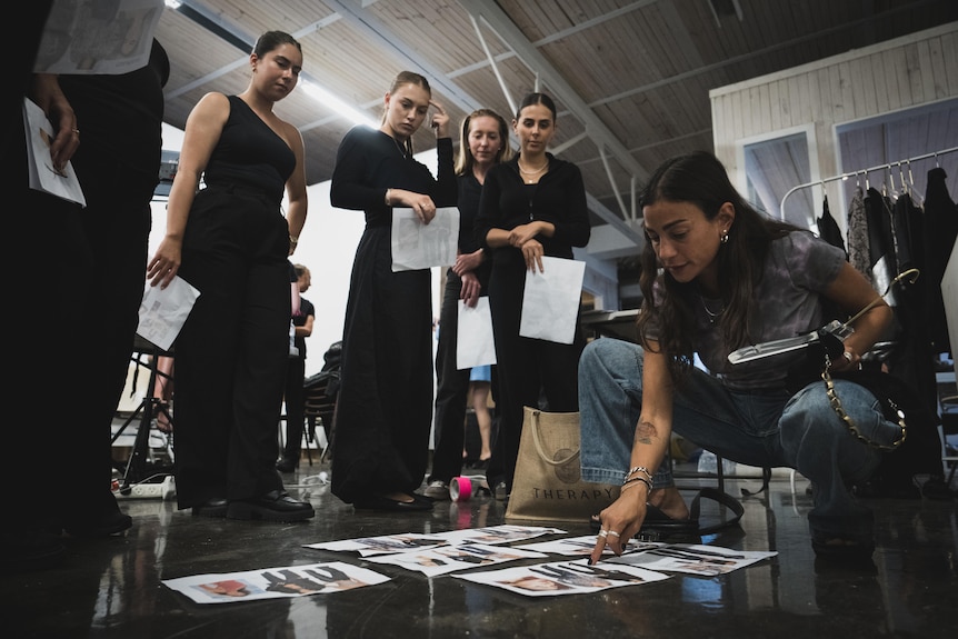 A woman squats in front of pages of paper with photos on them while a group of models watch over her shoulder.