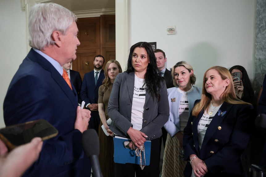 a male politician in a suit stands and talks to women who are survivors of epsteins abuse