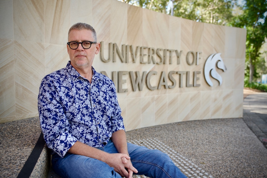 A man in jeans and a blue and white patterned shirt sits in front of a University of Newcastle sign.