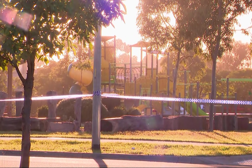 Blue and white police tape surrounds a park which contains a children's playground on a sunny morning.