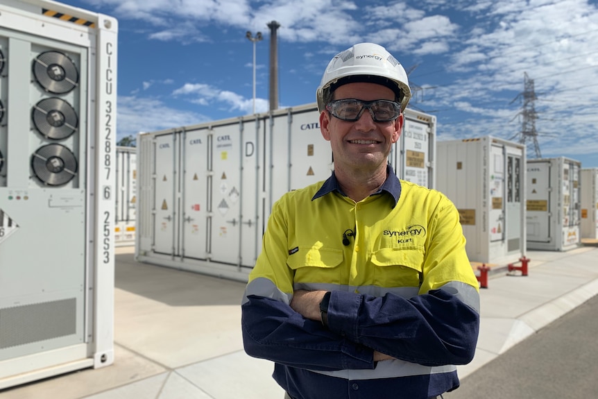 A man in high-vis wearing a hard hat stands before rows of white shipping-container-sized batteries