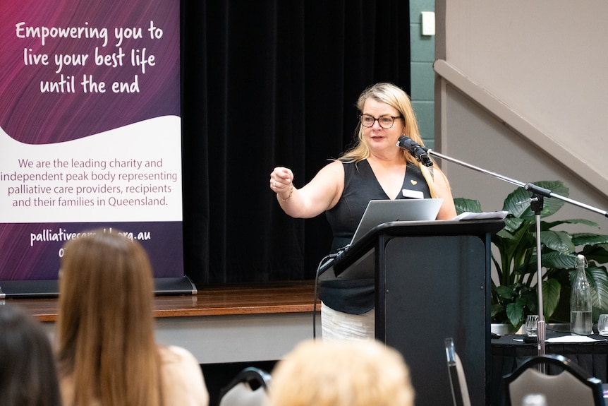 A woman wearing a black singlet and glasses stands at a lectern. She is pointing to a crowd as she talks.