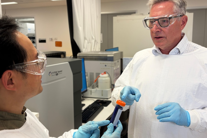 Two men in lab coats in a lab setting pass blood vials to each other.