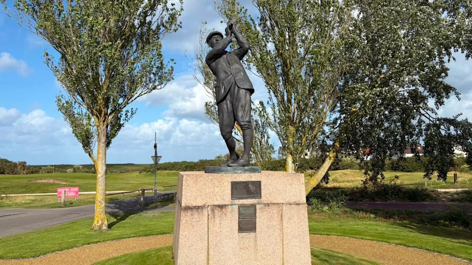 A large statue of a golfer (Harry Vardon) on a plinth outside the Royal Jersey Golf Club. The golf course is visible in the background.