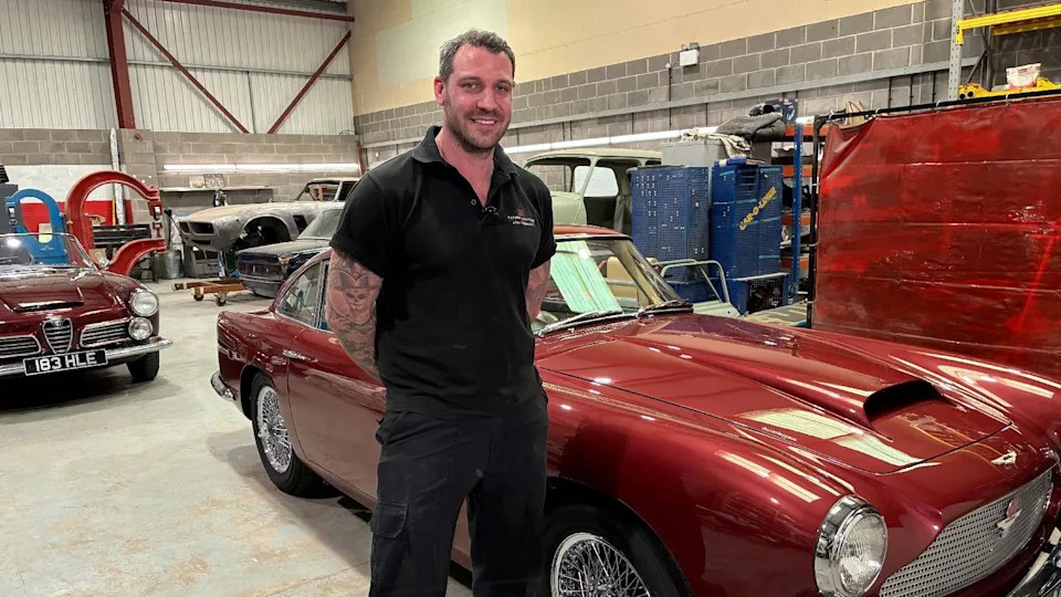 Alex Knight standing next to the 1961 Aston Martin DB4. He is smiling and has his hands behind his back. Alex has dark brown hair and a beard. He is wearing a black polo shirt and black trousers.