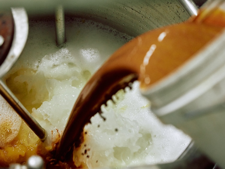 rusty coloured liquid is being poured from a bucket into a metal vat already filled with a milky substance