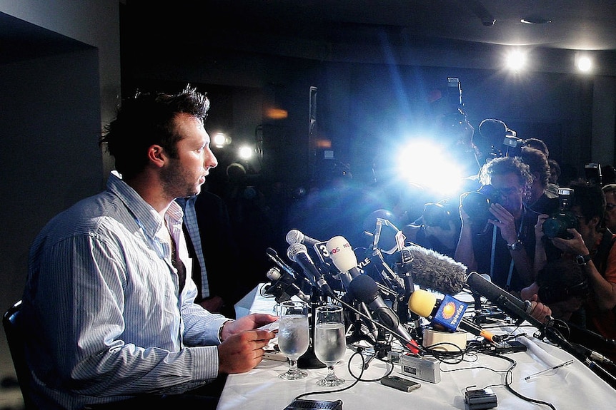Ian Thorpe surrounded by microphones and journalists as a bright flash goes off on a camera.