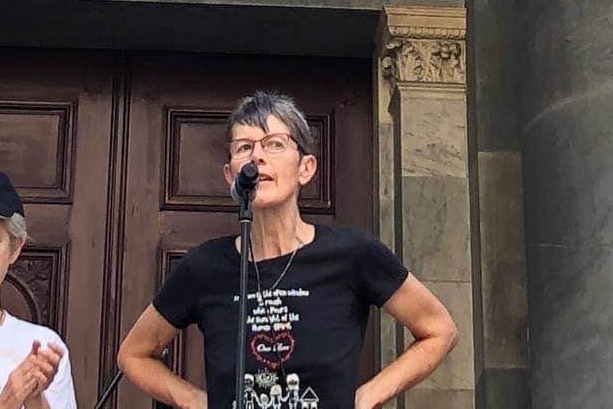 A woman speaking to a microphone with her hands on her hips between large grey columns