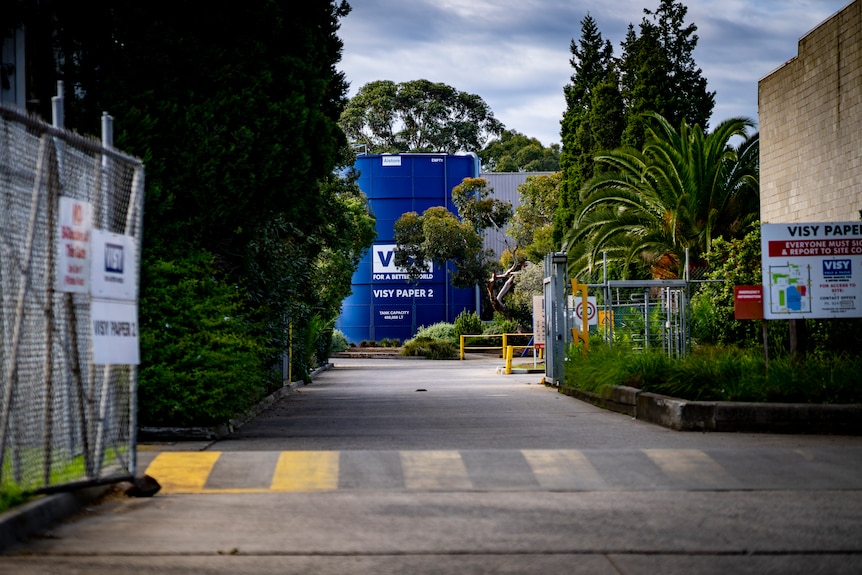 An open wire gate with "Visy" signs reveals a driveway lined with green trees leading to a blue tank that says "Visy Paper 2".