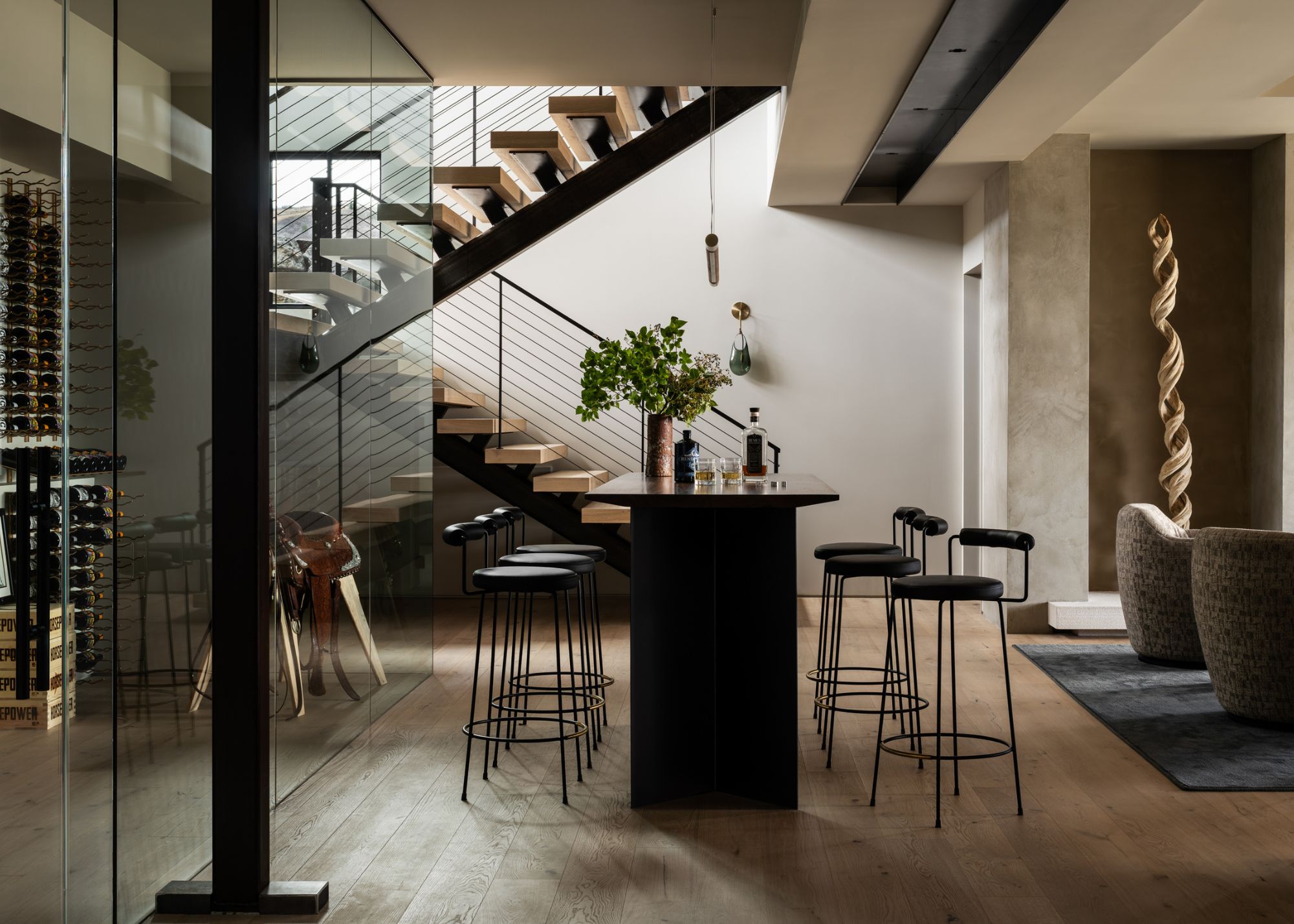 A contemporary wine room with floor-to-ceiling wine storage, a black island with stools on either side, with a staircase behind, and a petrified wood sculpture in a niche to the side of the space