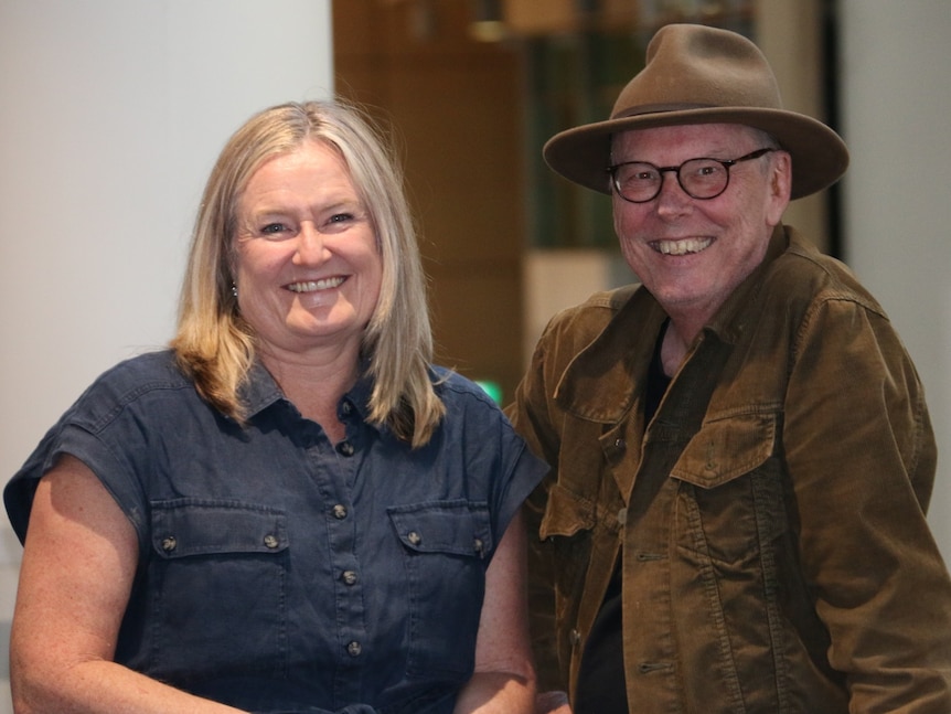 Blonde woman smiles big standing next to a man with thick-rimmed glasses and a brimmed hat