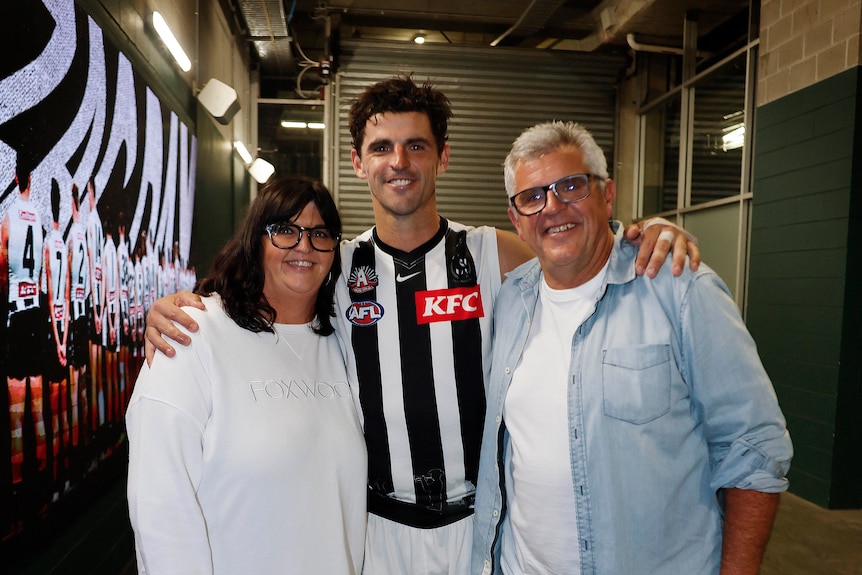 A smiling young AFL player in uniform stands with his arms around a dark-haired woman and white-haired man.
