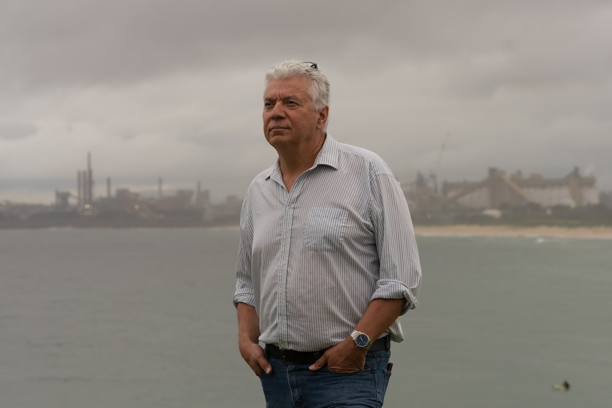 a man looking off in the distance with steelworks and water in the background