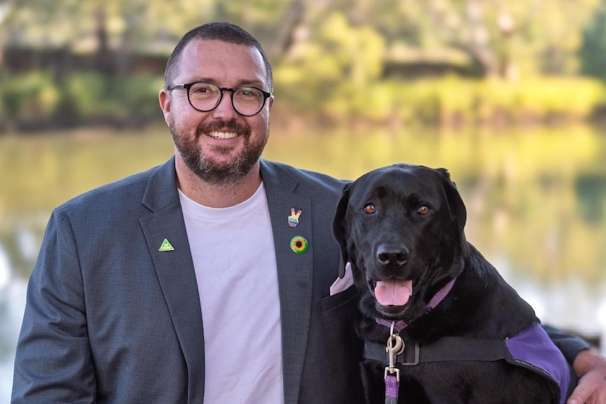 A man in a collared shirt and blazer wearing glasses pictured with a black dog in front of a river.