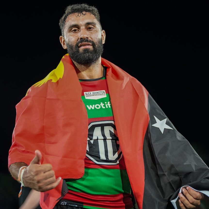 A Papua New Guinean man with a beard looks at the camera and wears a Papua New Guinea flag over his shoulders doing the shaka 