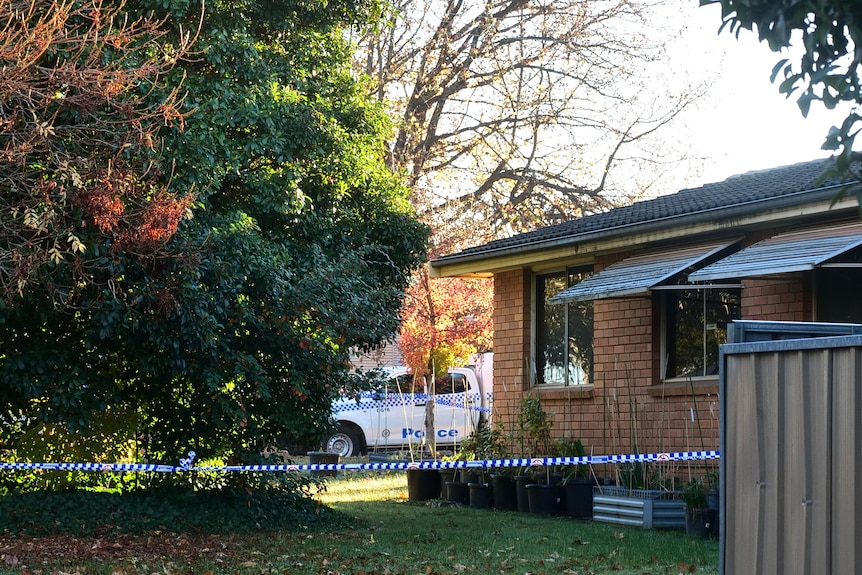Police tape around house, police car in distance. 