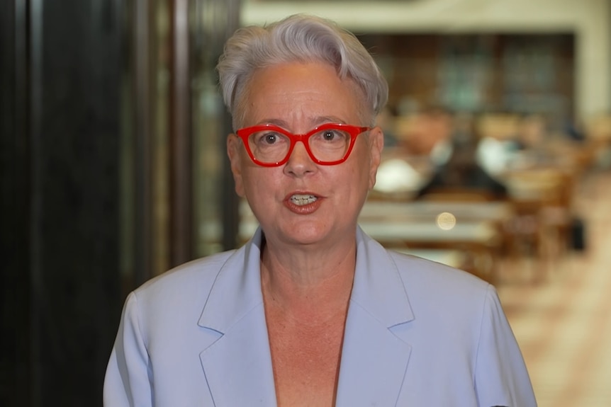 A woman with red-framed glasses stands behind a lectern as she talks to the media.
