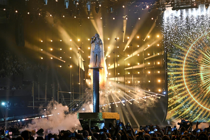 Delta Goodrem sings while standing on top of a tall podium coming out of a gold piano