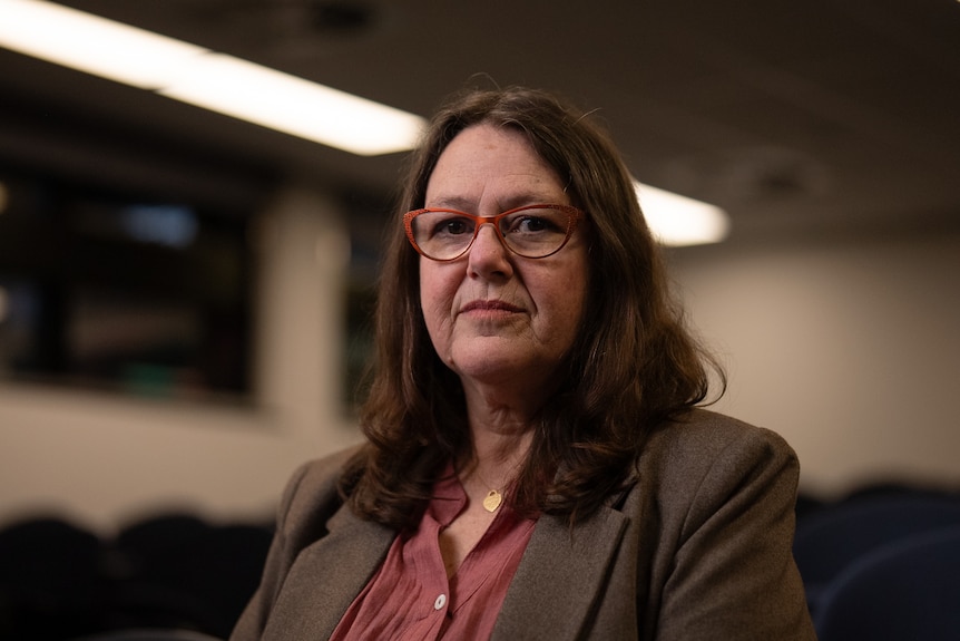A middle-aged woman wearing orange glasses, a brown blazer and rust blouse with long brown hair sitting in a lecture hall.