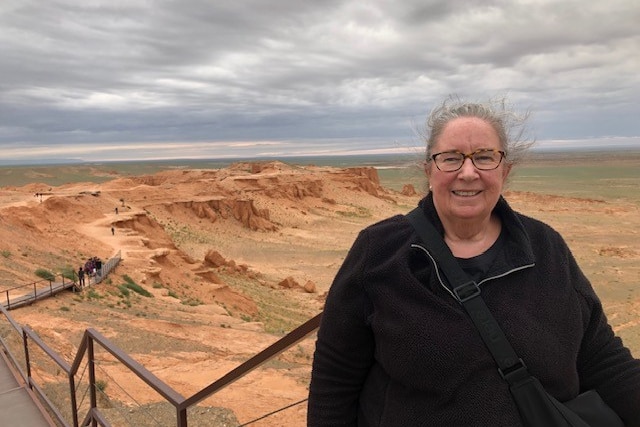 A woman stands on a long walking bridge with vast red dirt behind her.
