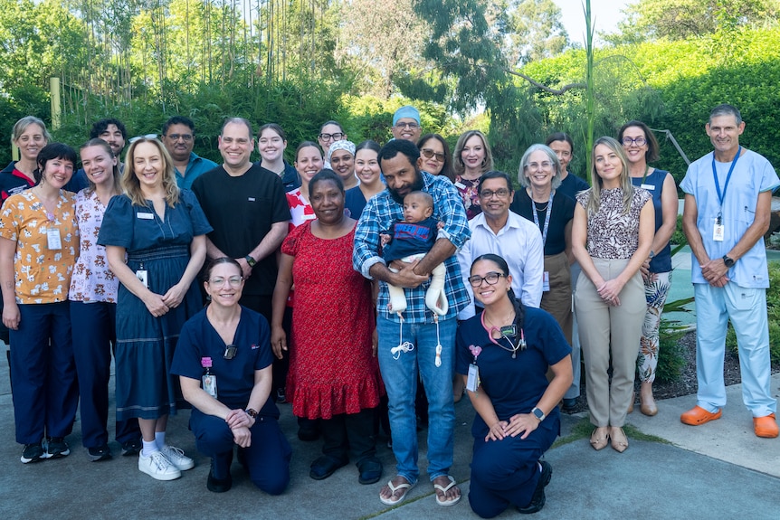 A Papua New Guinean mother and father pose with a baby boy and a team of medical staff on a parking lot.