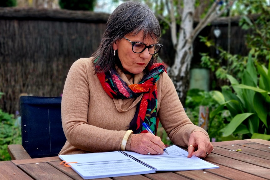 Brigid writes in her notebook on an outdoor table, surrounded by greenary.