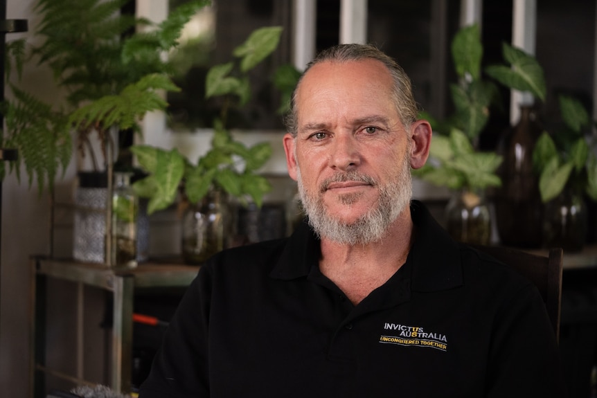 A bearded man wearing a black polo shirt smiling into the camera with plants in the background.
