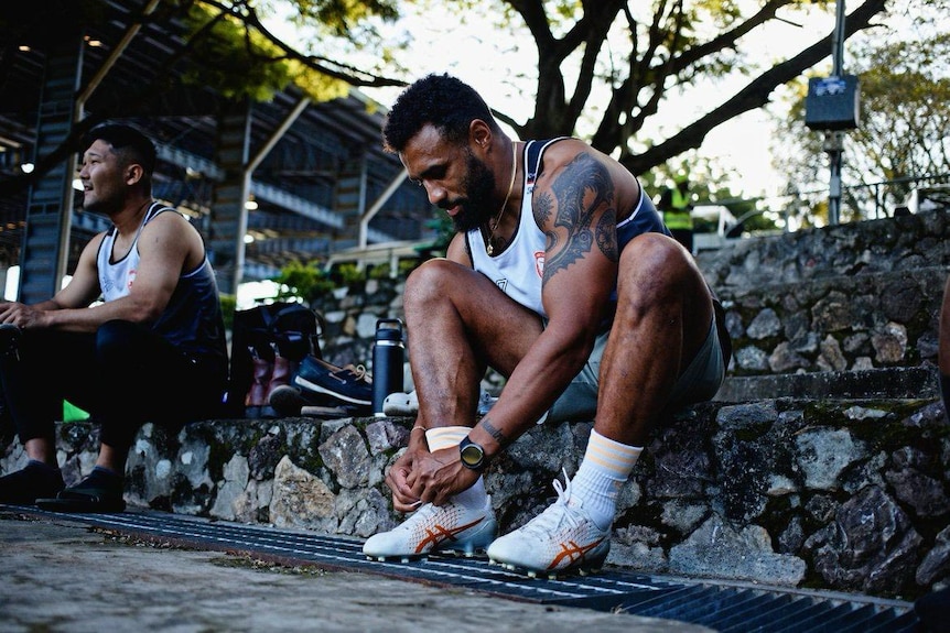 A Papua New Guinea man sits down on stone steps wearing rugby training gear and ties his rugby boots laces