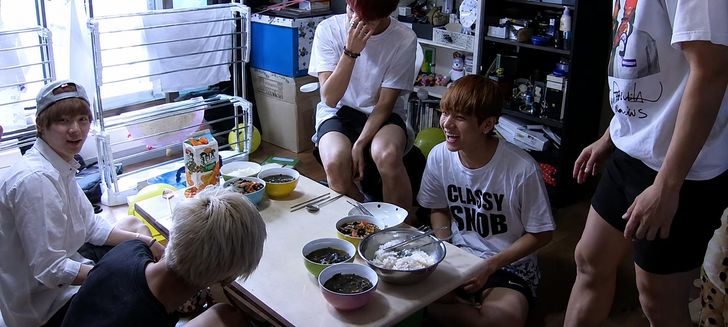The seven members of BTS lived together in one home as trainees, sharing a single bathroom. The members prepare a meal together in a living room crowded with household items, including a drying rack. Courtesy of BigHit Music