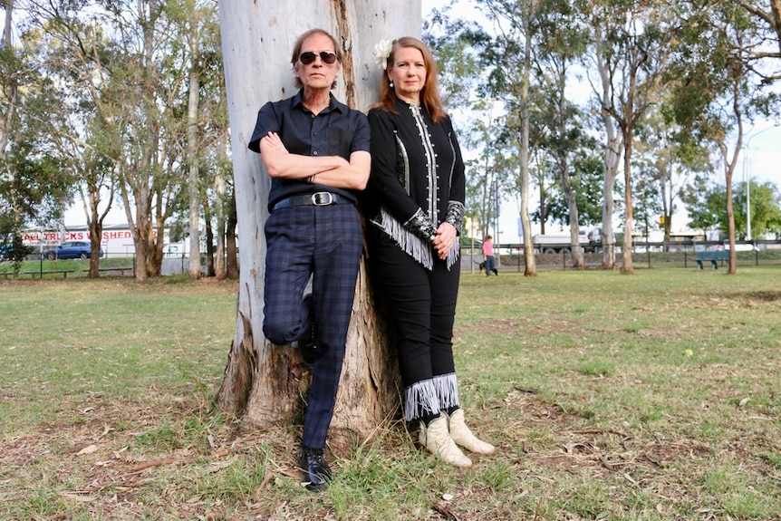 A man in his 60s with blue and black plaid pants and a black stands and a woman in a tasseled outfit stand against a tree