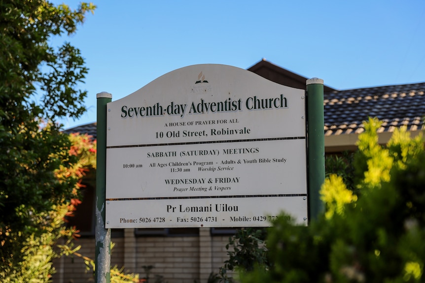 A sign outside a building reads: Seventh-day Adventist Church' there are trees either side of the sign.