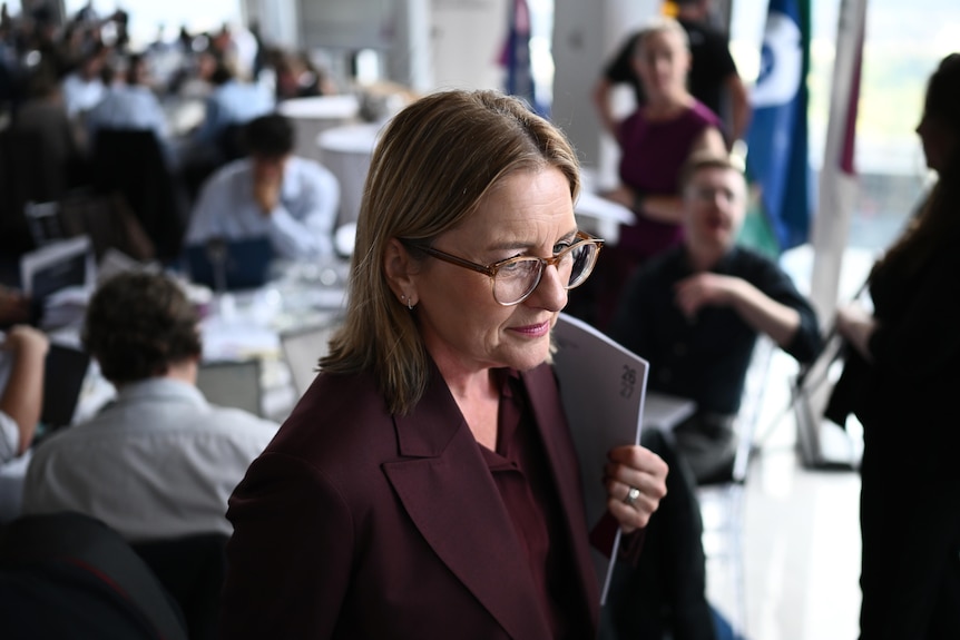 A woman wearing glasses holds a document while speaking to reporters in a conference room.