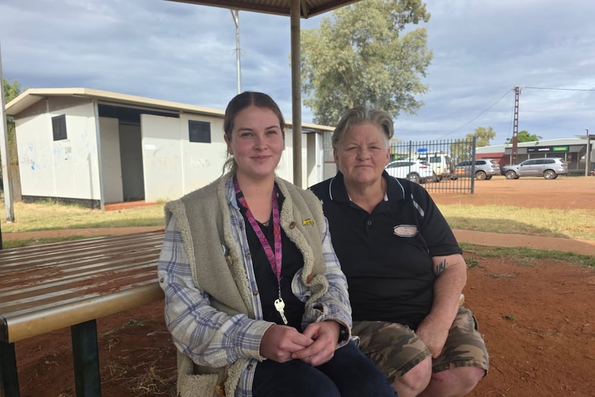 Two women sit on park bench in front of table in remote setting with red dirt