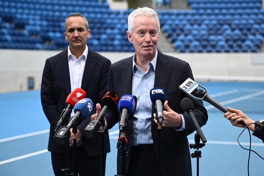 Two men in suits, no tie, standing in front of microphone, press conference, on a tennis court