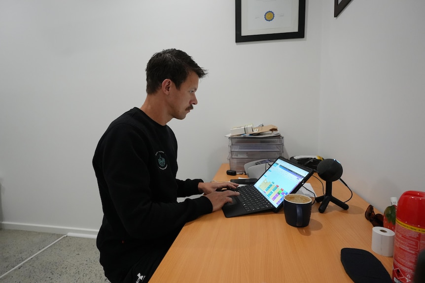 A man sits at a desk and looks at a laptop.