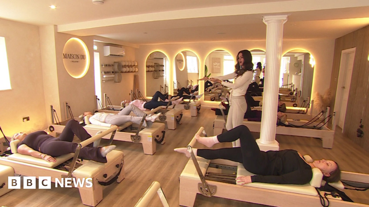 A Reformer Pilates studio with seven machines, each one being used by a woman, and a female instructor stood in the middle of the room