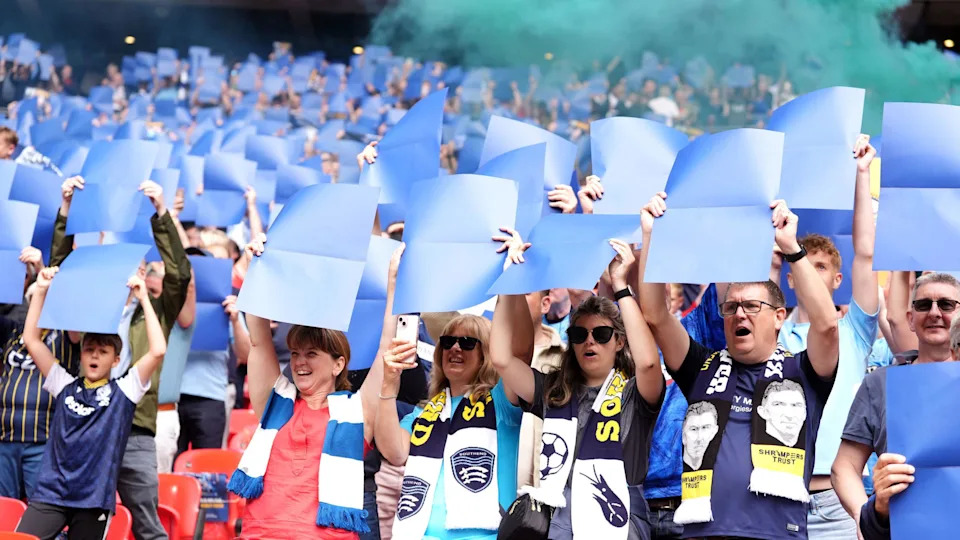 Southend supporters stand up in their seats at Wembley stadium, holding large blue sheets of paper above their heads. Many wear their club's scarves and kit. There is blue smoke wafting above them.