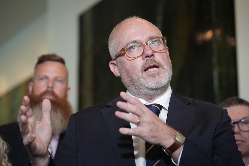 Tim Ayres in mid talk, bald, Caucasian, grey stubble, wears a black suit and tie.