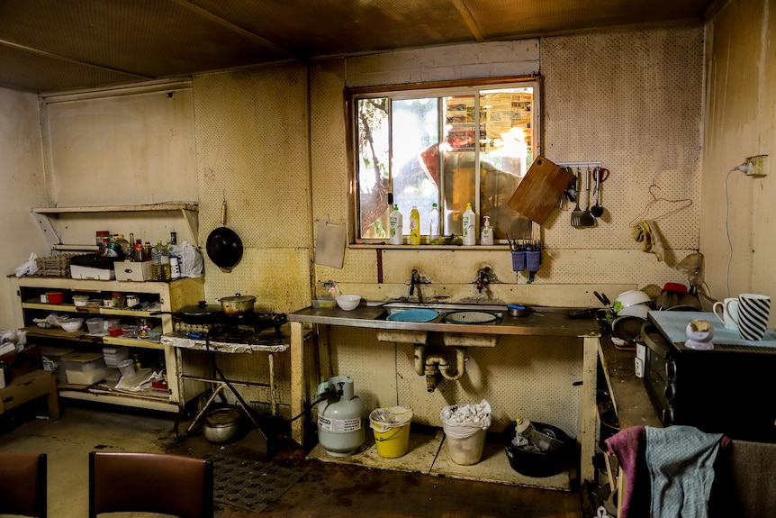 A dirty kitchen with a gas bottle connected to a cooktop and buckets in sink beneath a peeling cracked wall and windows