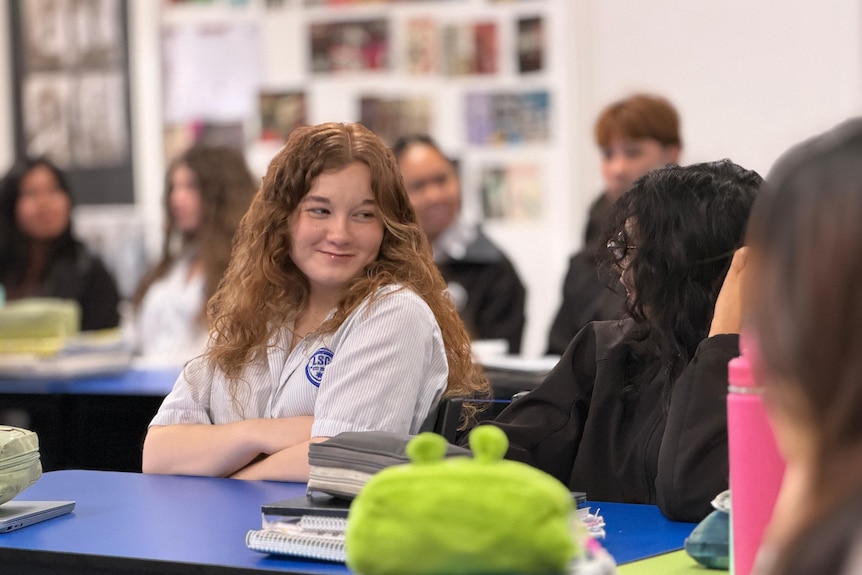 A girl with long, wavy hair smiles at a dark-haired girl while they sit in a classroom.