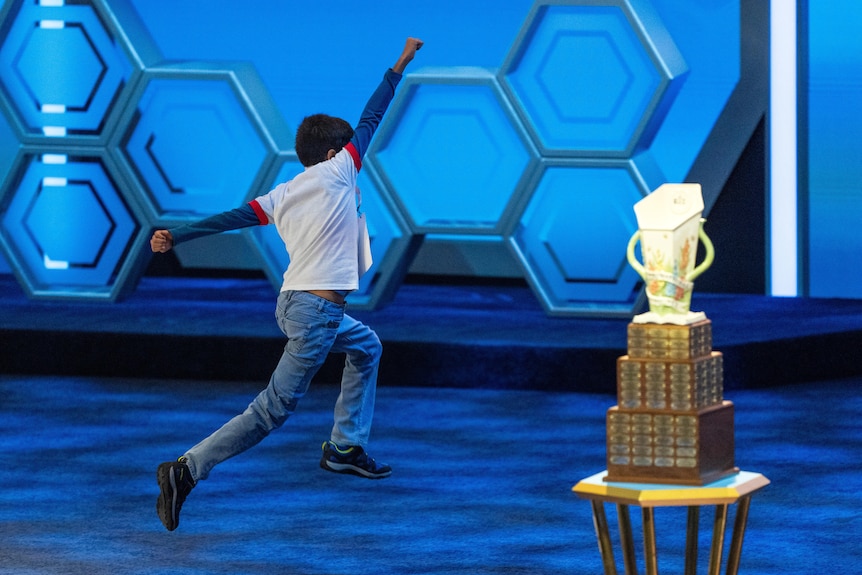 A boy bounds off the stage with one arm in the air, next to a trophy on display